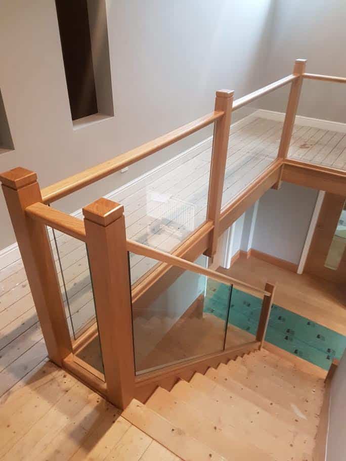 View down an oak staircase with square wooden newel posts, matching handrails and clear glass panels enclosing the staircase and upper landing.