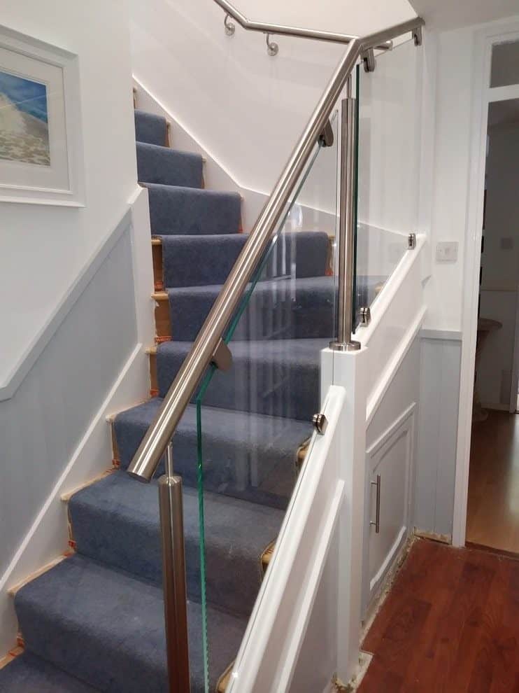 Blue carpeted staircase with clear clamped glass panels, stainless steel posts and handrails above white panelling and an understair cupboard.