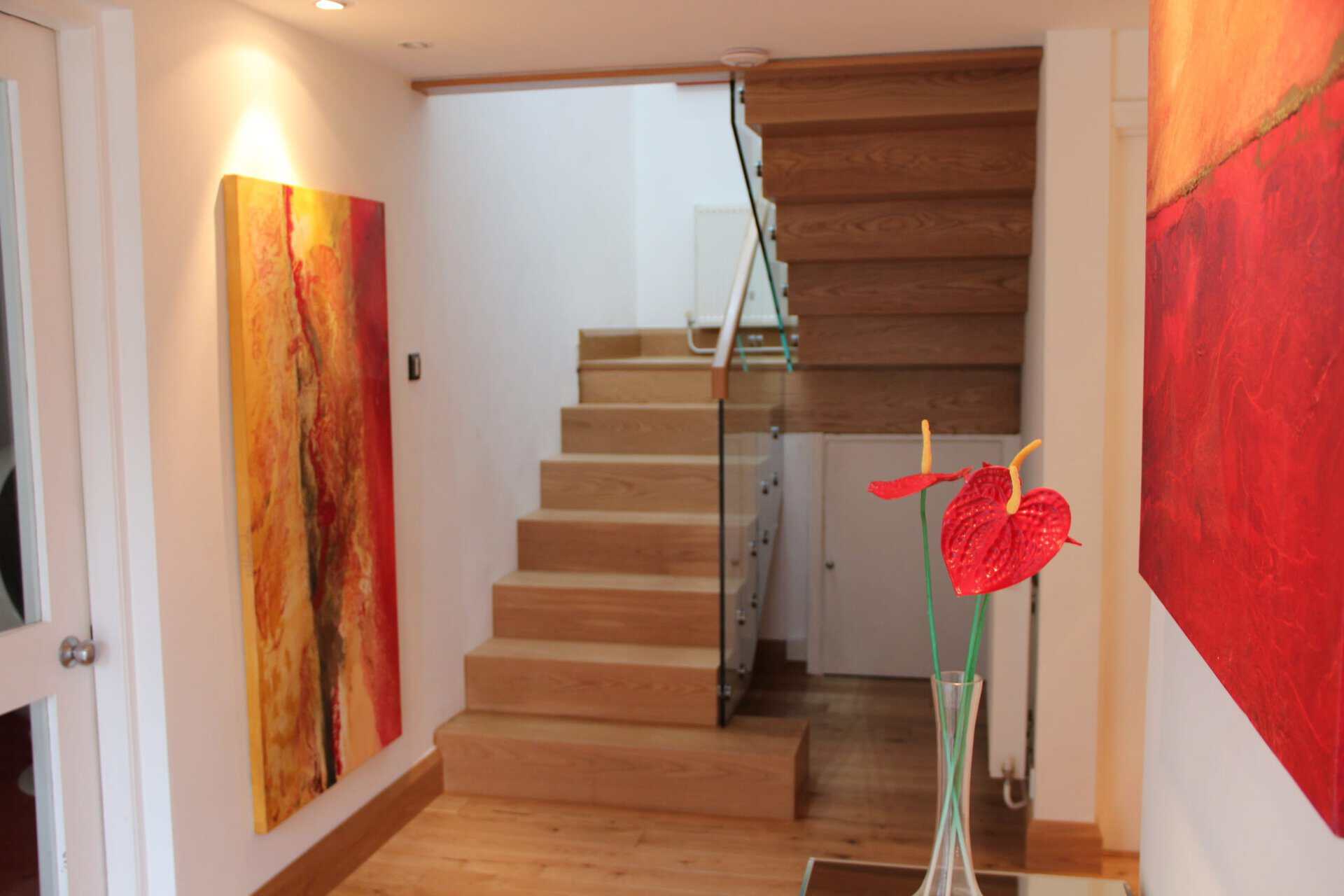 Turning oak staircase with closed risers, glass balustrades and a metal handrail in a white hallway with red abstract artwork.
