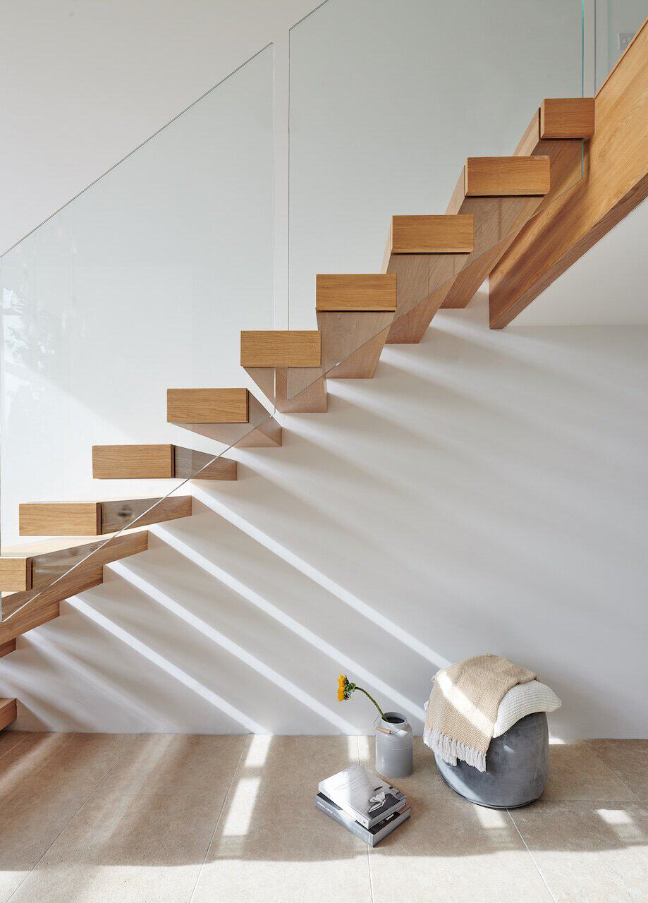 Side view of an open-riser oak staircase with a zigzag stringer and frameless glass balustrade casting shadows on a white wall.
