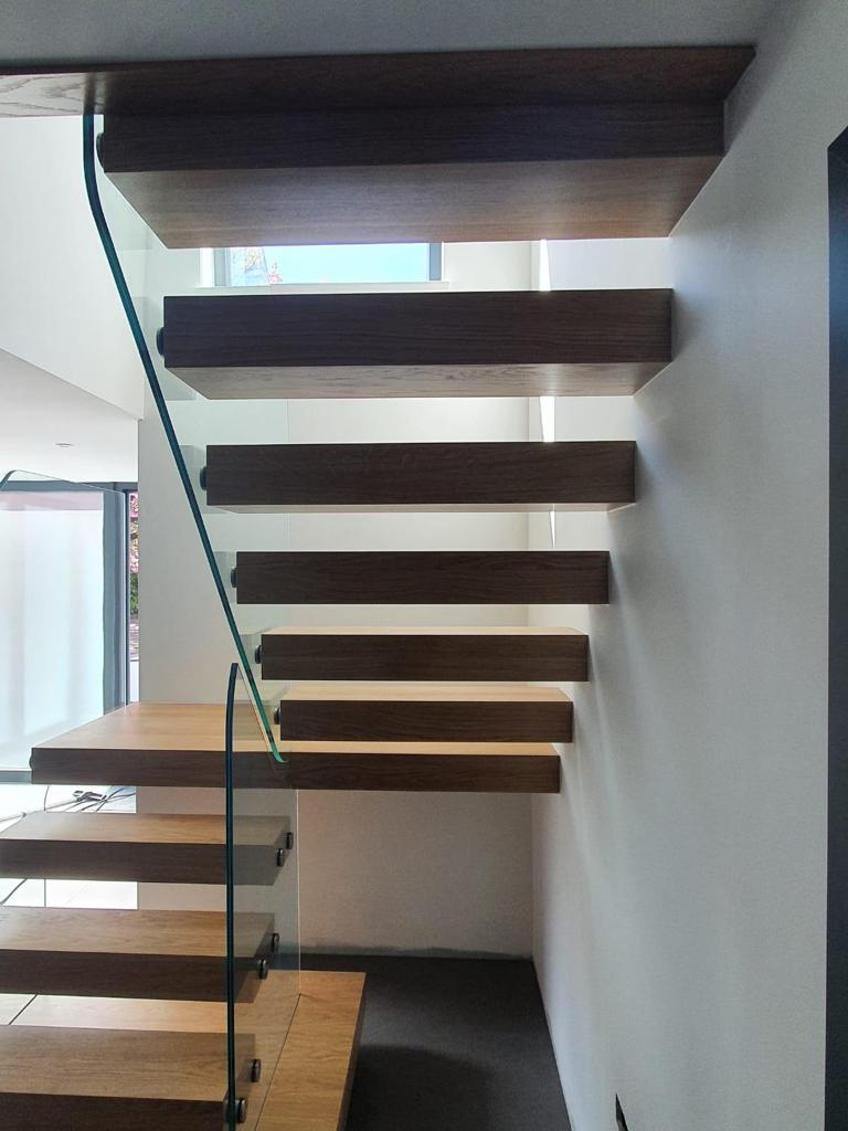 Close view of thick wooden floating stair treads with open risers and a frameless glass balustrade in the Rolling family home.