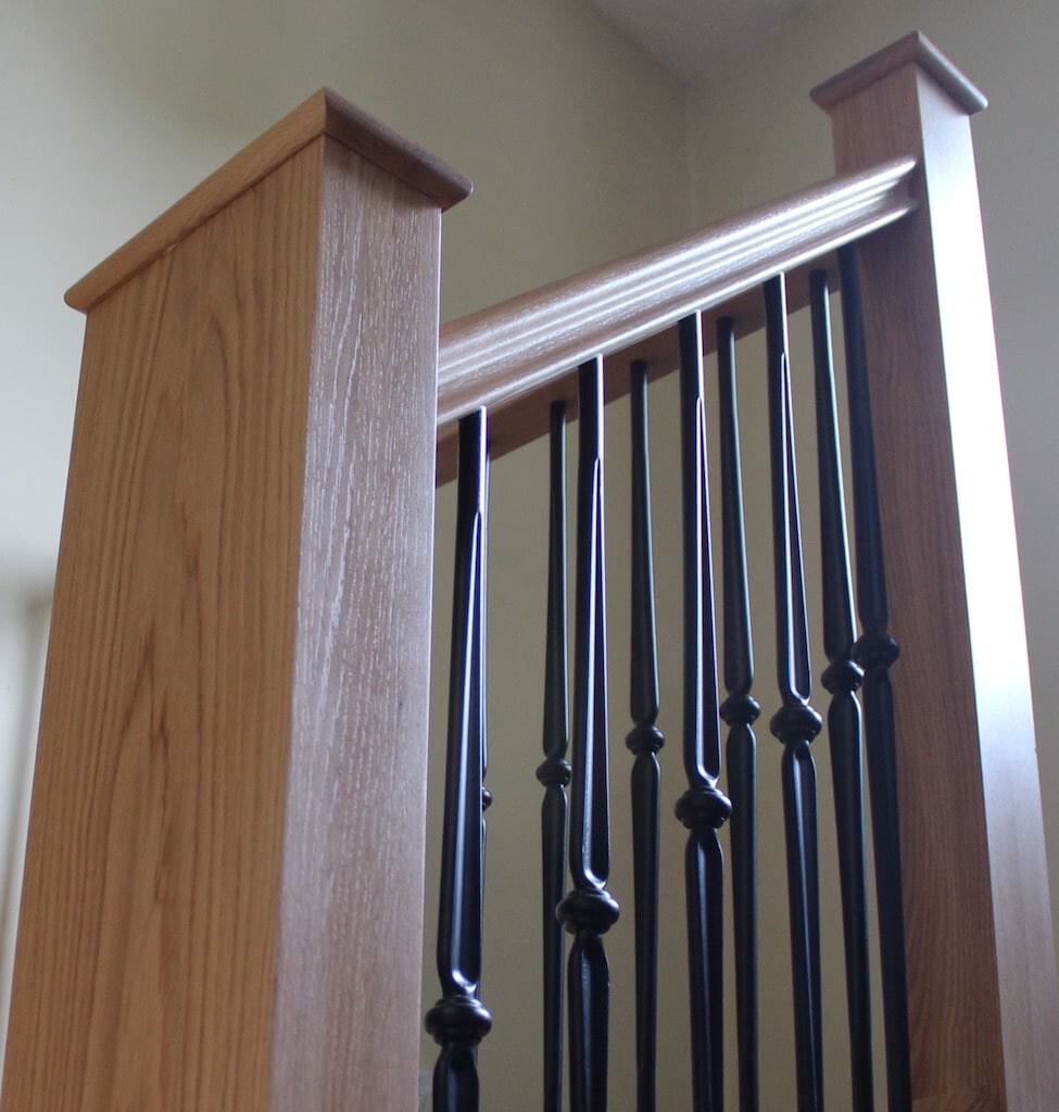 Close-up of decorative black metal spindles between square oak newel posts beneath an oak handrail.
