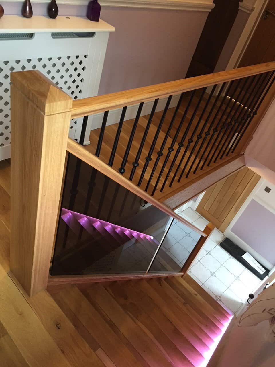 View down an oak staircase with glass panels, purple step lighting and black decorative metal spindles along the upper landing.