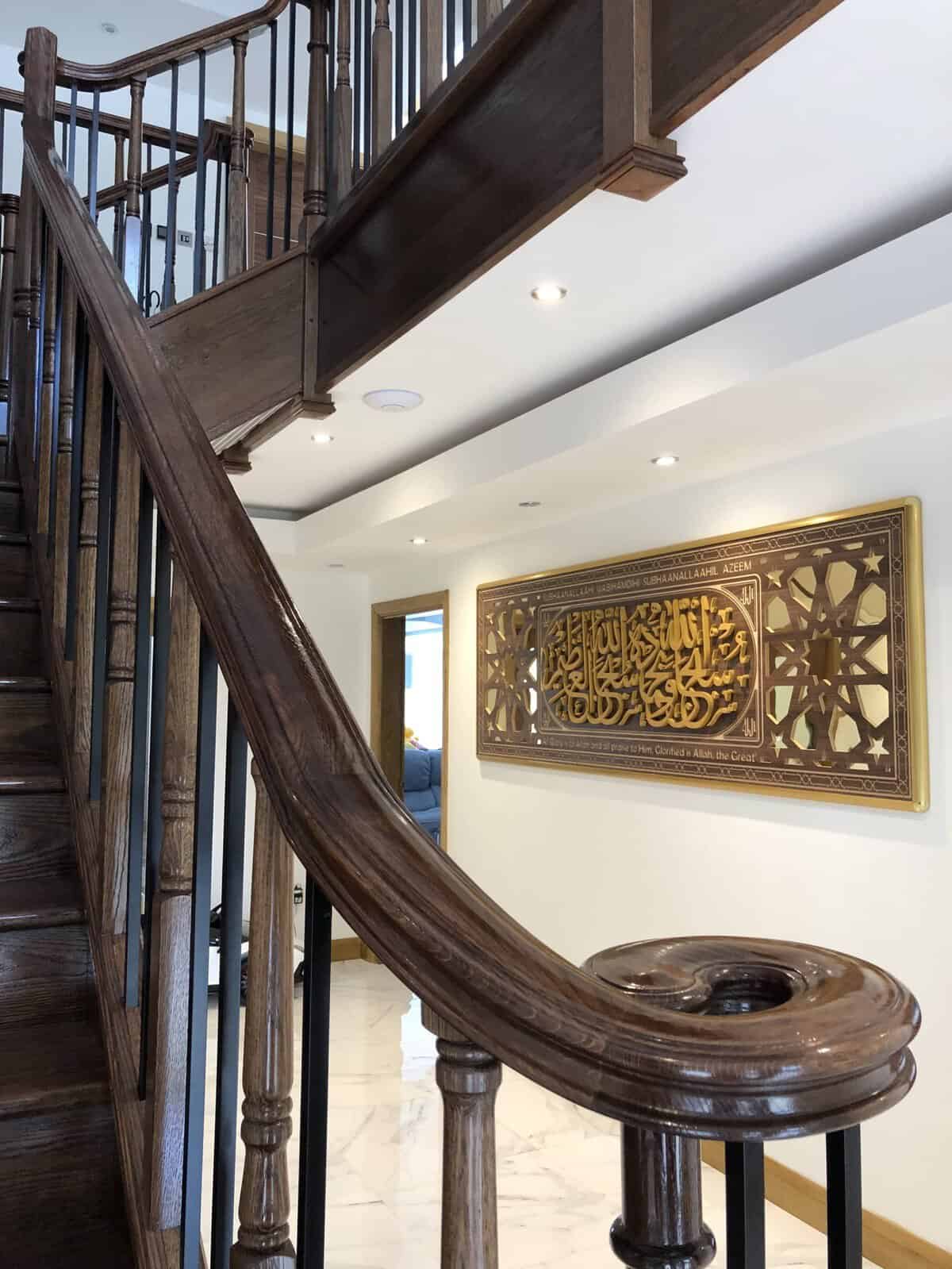 Close-up of a dark wooden staircase handrail ending in a carved scroll above decorative metal spindles, with a matching gallery landing overhead.