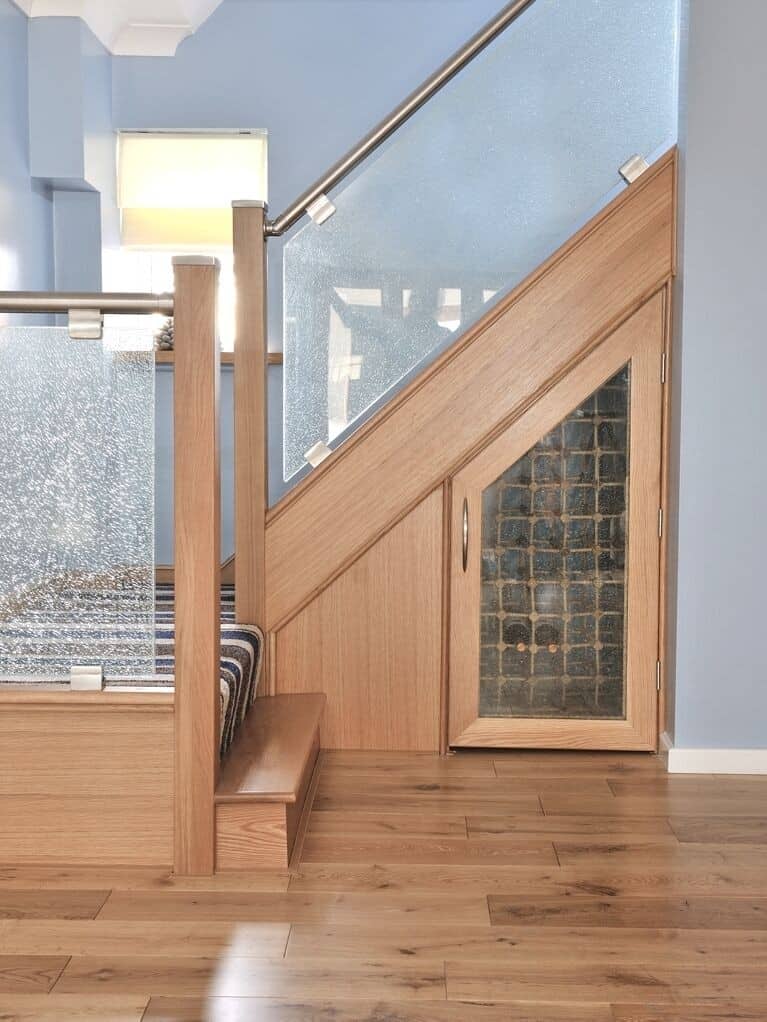 Fitted oak cupboard with a patterned glass door beneath a staircase with glass balustrades and a metal handrail.