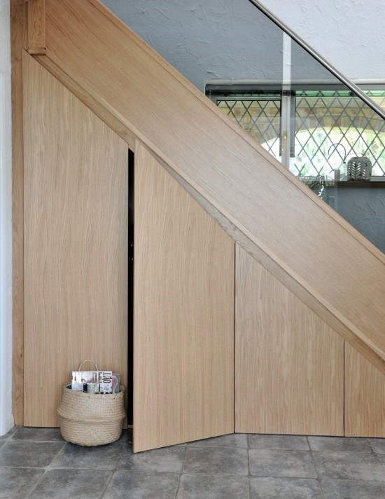 Flush oak cupboard doors fitted beneath a staircase, following its sloping profile, with one door partly open.